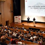 Dwaj mężczyźni na scenie. Przed nimi pełna widownia. W tle ekran na scenie z wyświetlonymi slajdami.