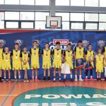 Zawody koszykarskie na hali sportowej. Grupowe zdjęcie zwycięskiej drużyny. Młodzi chłopcy stoją w szeregu, z medalami zawieszonymi na szyjach, i pozują do zdjęcia. Po obu ich stronach stoją też dorośli: ich opiekunowie i VIP-y.