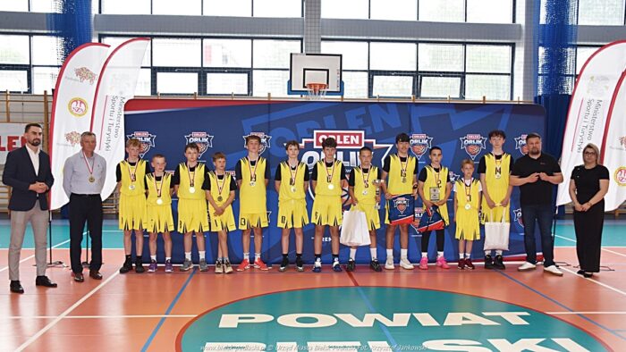 Zawody koszykarskie na hali sportowej. Grupowe zdjęcie zwycięskiej drużyny. Młodzi chłopcy stoją w szeregu, z medalami zawieszonymi na szyjach, i pozują do zdjęcia. Po obu ich stronach stoją też dorośli: ich opiekunowie i VIP-y.