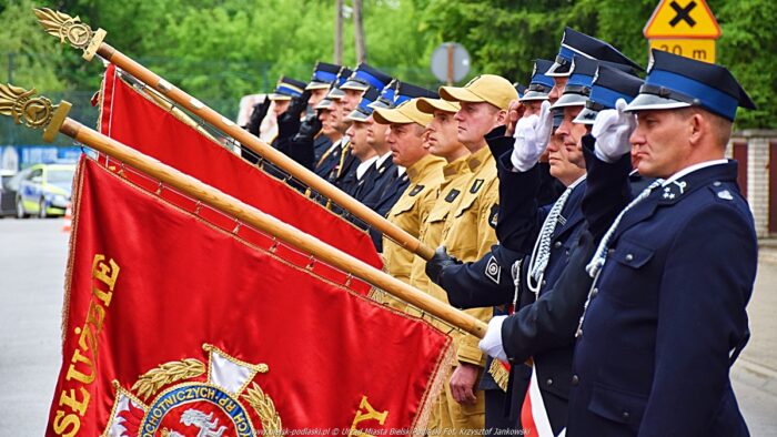 Uroczystości przed komendą. Strażacy ustawieni w dwuszeregu, w odświętnych mundurach, prezentują kolorowe chorągwie. W tle zieleń miejska.
