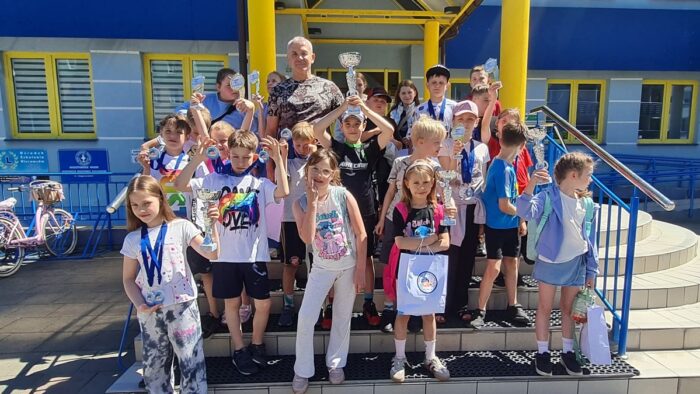 Grupa kolorowy ubranych dzieci radośnie pozuje do zdjęcia, stojąc na schodach pływalni.