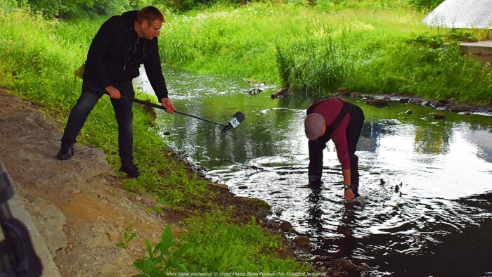 Dwaj mężczyźni pod mostem nad rzeką. Dziennikarz stoi na brzegu i trzyma mikrofon na wysięgniku. W wodzie stoi drugi mężczyzna i nabiera brudnej wody do szklanego pojemnika.
