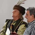 Dwaj mężczyźni przemawiają. Mężczyzna z lewej ubrany jest w strój szlachcica.