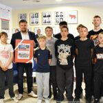 Grupowe zdjęcie koszykarzy, trenera i burmistrzów. Wspólnie pozują do zdjęcia stojąc na korytarzu urzędu. Burmistrz trzyma ramę ze zdjęciami zespołu z ostatnich turniejów.
