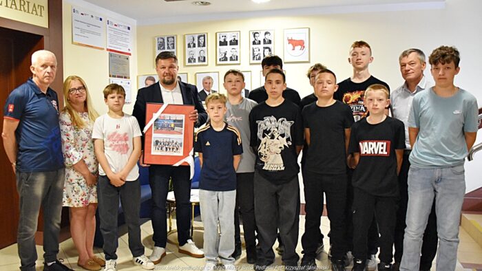 Grupowe zdjęcie koszykarzy, trenera i burmistrzów. Wspólnie pozują do zdjęcia stojąc na korytarzu urzędu. Burmistrz trzyma ramę ze zdjęciami zespołu z ostatnich turniejów.