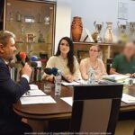 Grupa osób dorosłych dyskutuje przy stole. Z lewej mężczyzna mówi do mikrofonu. Przed nim kilka osób siedzi za stołem i go słucha.