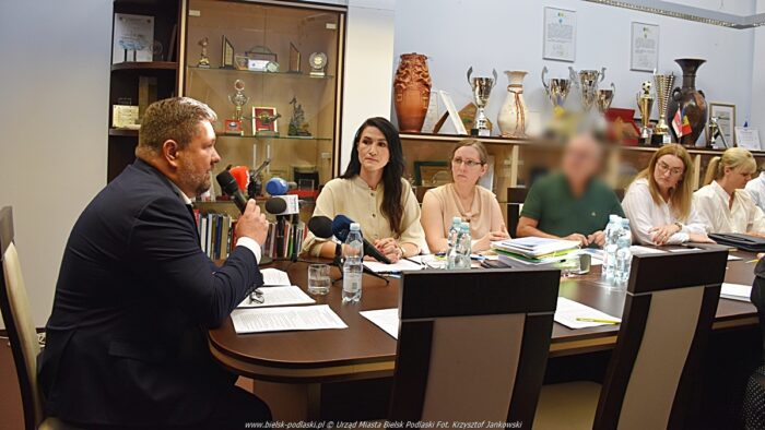 Grupa osób dorosłych dyskutuje przy stole. Z lewej mężczyzna mówi do mikrofonu. Przed nim kilka osób siedzi za stołem i go słucha.