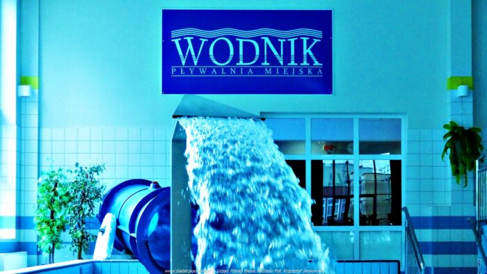 Wnętrze pływalni krytej. Na ścianie szyld z napisem "Wodnik". Przed nim urządzenie nalewające wodę do niecki brodziku.