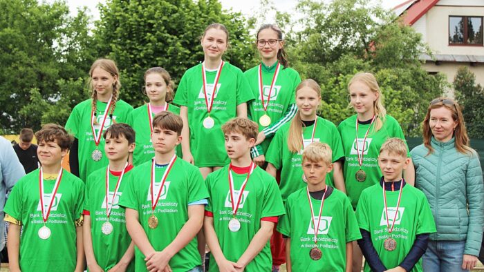 Młodzi lekkoatleci podczas zawodów i wręczania nagród. Kilkanaście zawodniczek w zielonych strojach z logo sponsora zawodów stoi w trzech szeregach na podium.