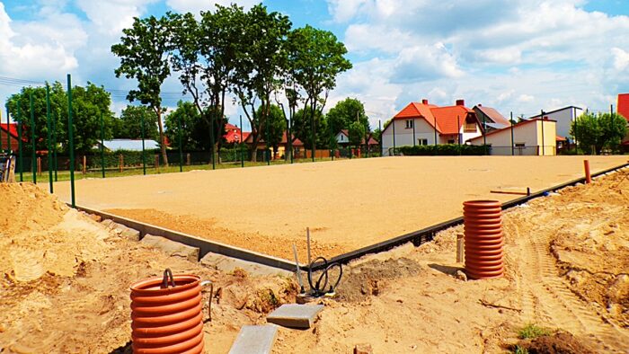Budowa boiska do siatkówki plażowej. Przestrzeń pokryta piachem, ustawione krawężniki, w okół sprzęt budowany, w tle zabudowania osiedla jednorodzinnego i zieleń drzew.