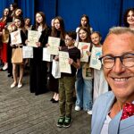 Poziome zdjęcie laureatów nagród festiwalowych na drewnianej scenie muszli koncertowej. Na pierwszym planie mężczyzna, który robi selfie z ustawionymi za nimi młodymi artystami.