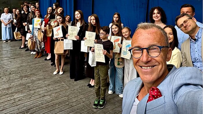Poziome zdjęcie laureatów nagród festiwalowych na drewnianej scenie muszli koncertowej. Na pierwszym planie mężczyzna, który robi selfie z ustawionymi za nimi młodymi artystami.