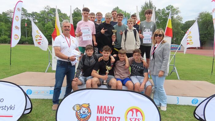 Grupa zawodników z trenerem stojąca na podium na tle stadionu lekkoatletycznego.