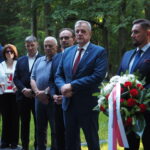 zdjęcie przedstawia mężczyzn i kobietę stojących w rzędzie, pierwszy trzyma biało niebieski wieniec
