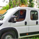 Zdjęcie samochodu dostawczego na parkingu, wśród miejskiej zieleni. Przez okno samochodu widać twarz kierowcy.