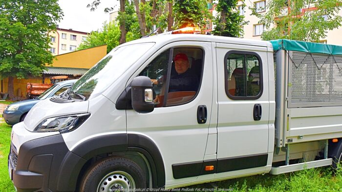 Zdjęcie samochodu dostawczego na parkingu, wśród miejskiej zieleni. Przez okno samochodu widać twarz kierowcy.
