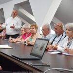 Kilkanaście osób dorosłych siedzi za stołem podczas spotkania informacyjnego.