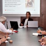 Kobieta prowadzi prezentację dla kilkunastu osób w sali konferencyjnej.