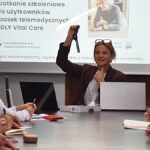 Kobieta prowadzi prezentację dla kilkunastu osób w sali konferencyjnej.