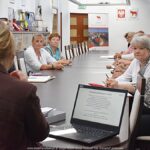 Kilkanaście osób dorosłych siedzi za stołem podczas spotkania informacyjnego.