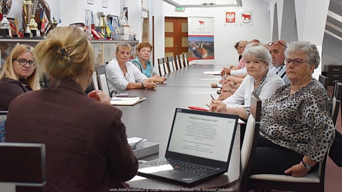 Kilkanaście osób dorosłych siedzi za stołem podczas spotkania informacyjnego.
