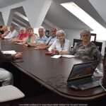 Kilkanaście osób dorosłych siedzi za stołem podczas spotkania informacyjnego.